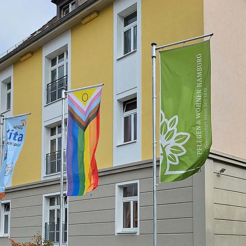 Gebäude von PFLEGEN & WOHNEN HAMBURG mit Fahnenmasten, an denen eine Regenbogenflagge für Diversity weht.