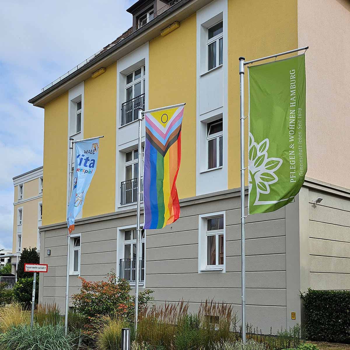 Gebäude von PFLEGEN & WOHNEN HAMBURG mit Fahnenmasten, an denen eine Regenbogenflagge für Diversity weht.